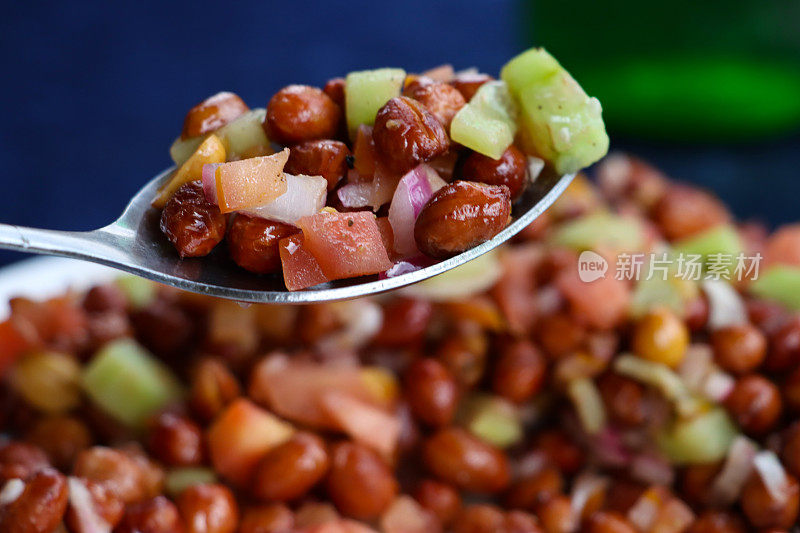 特写图片一勺自制的花生masala chaat，白碗的街头风味小吃，花生，chaat masala，辣椒粉，香菜，serrano辣椒片(Capsicum annuum)，番茄丁和红洋葱，蓝色背景，重点在前景